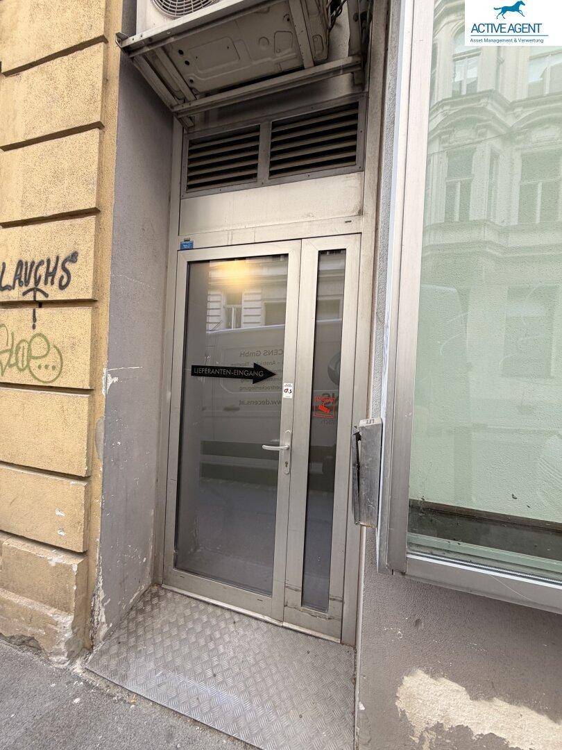 Nebeneingang mit Metallrampe und Lüftungseinheit über der Tür, an einer Gebäudeseite gelegen.