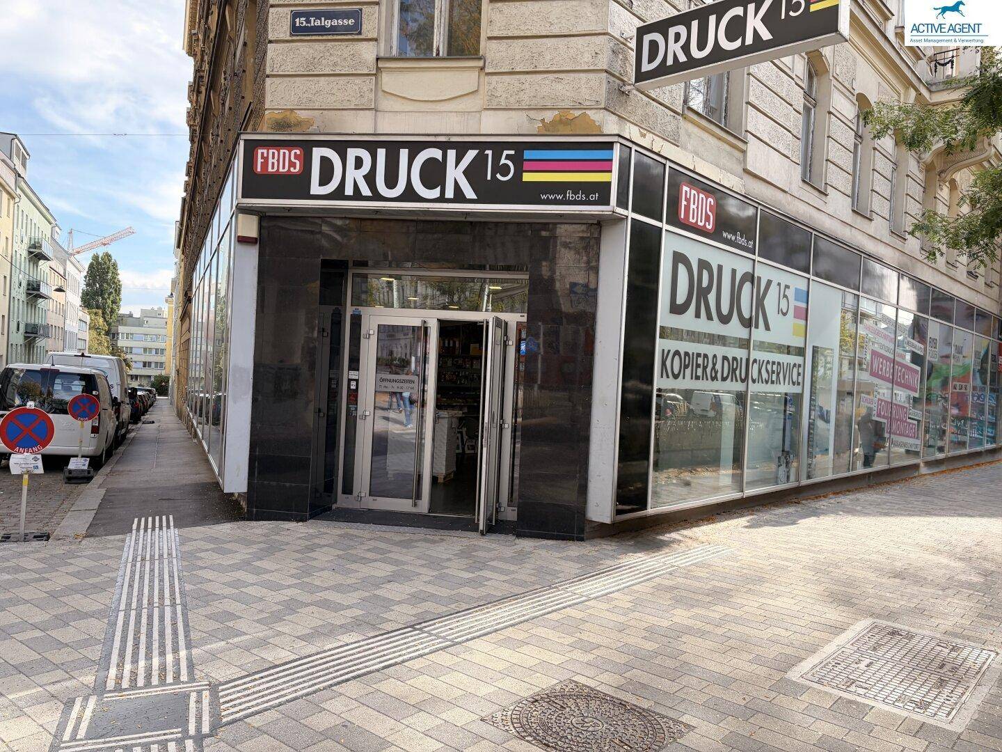 Eingangsbereich des Geschäfts an einer Straßenecke mit großen Schaufenstern und Bürgersteig.