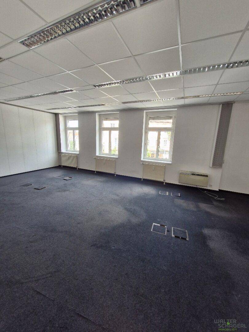 Großer, offener Büroraum mit dunklem Teppichboden, vielen Fenstern und abgehängter Decke, bereit zur Nutzung.