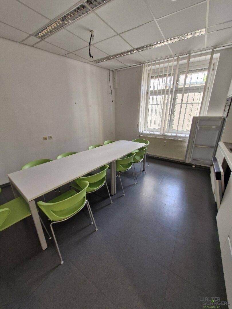 Funktionale Teeküche mit langem Tisch, grünen Stühlen und dunklem Vinylboden, ideal für Pausen.