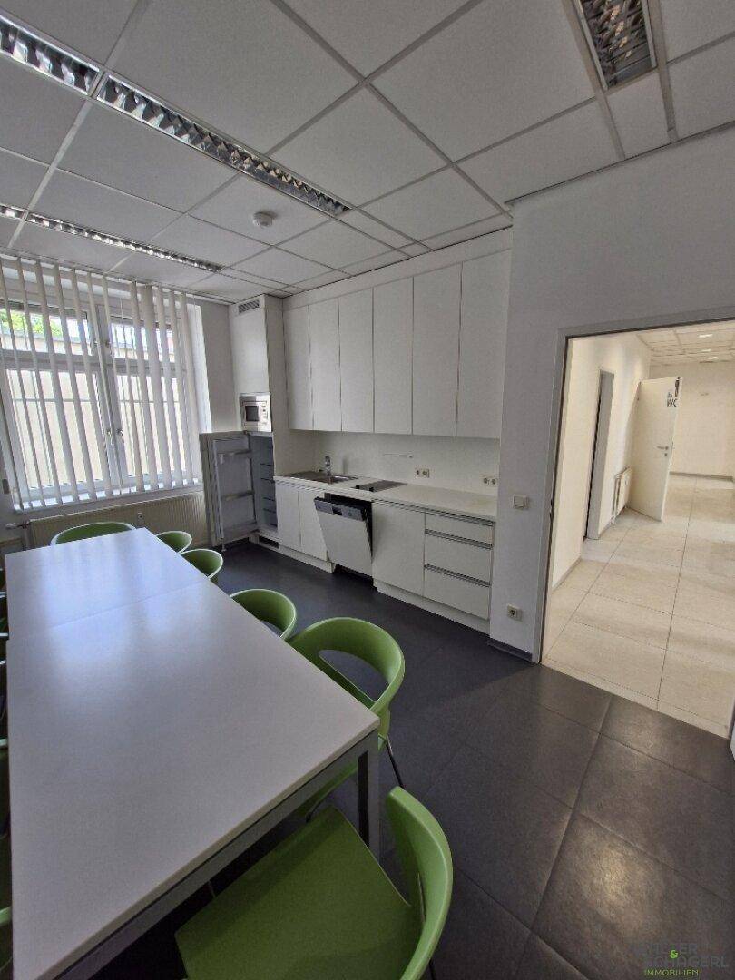 Helle Teeküche mit weißen Schränken, Spüle, Geschirrspüler und Kühlschrank, neben einem langen Esstisch.