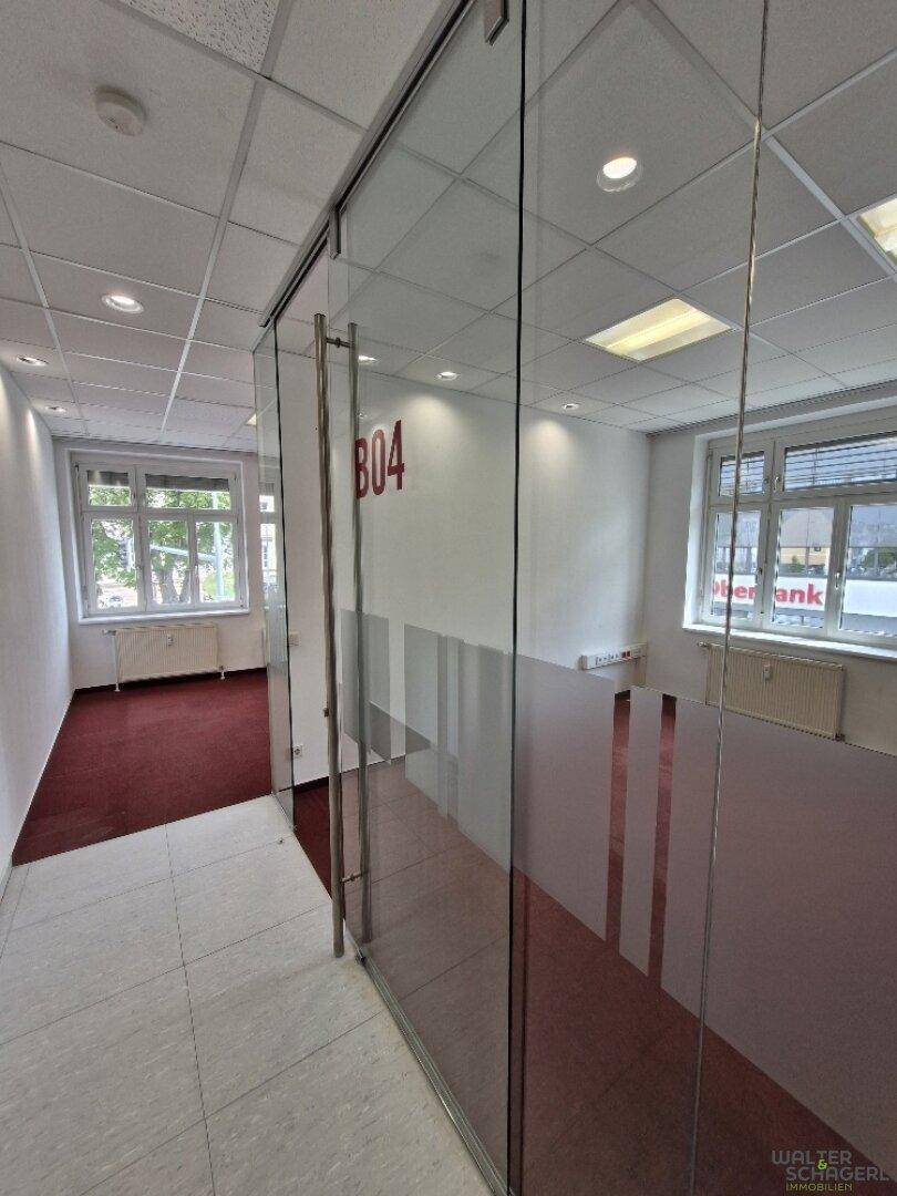 Moderner Flur mit Glaswänden und Fliesenboden, der zu einem Büro mit rotem Teppich führt.