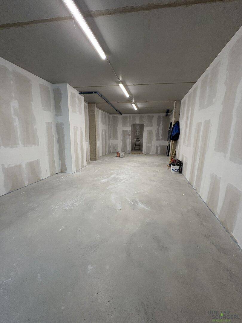 Großer, leerer Raum im Rohbau mit Trockenbauwänden und Betonboden, beleuchtet von Deckenleuchten.