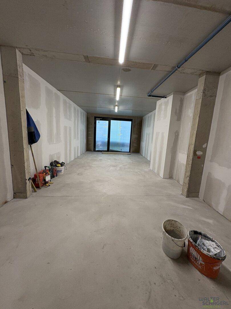 Langer Rohbauraum mit Betonboden und Trockenbauwänden, am Ende eine Tür oder großes Fenster.
