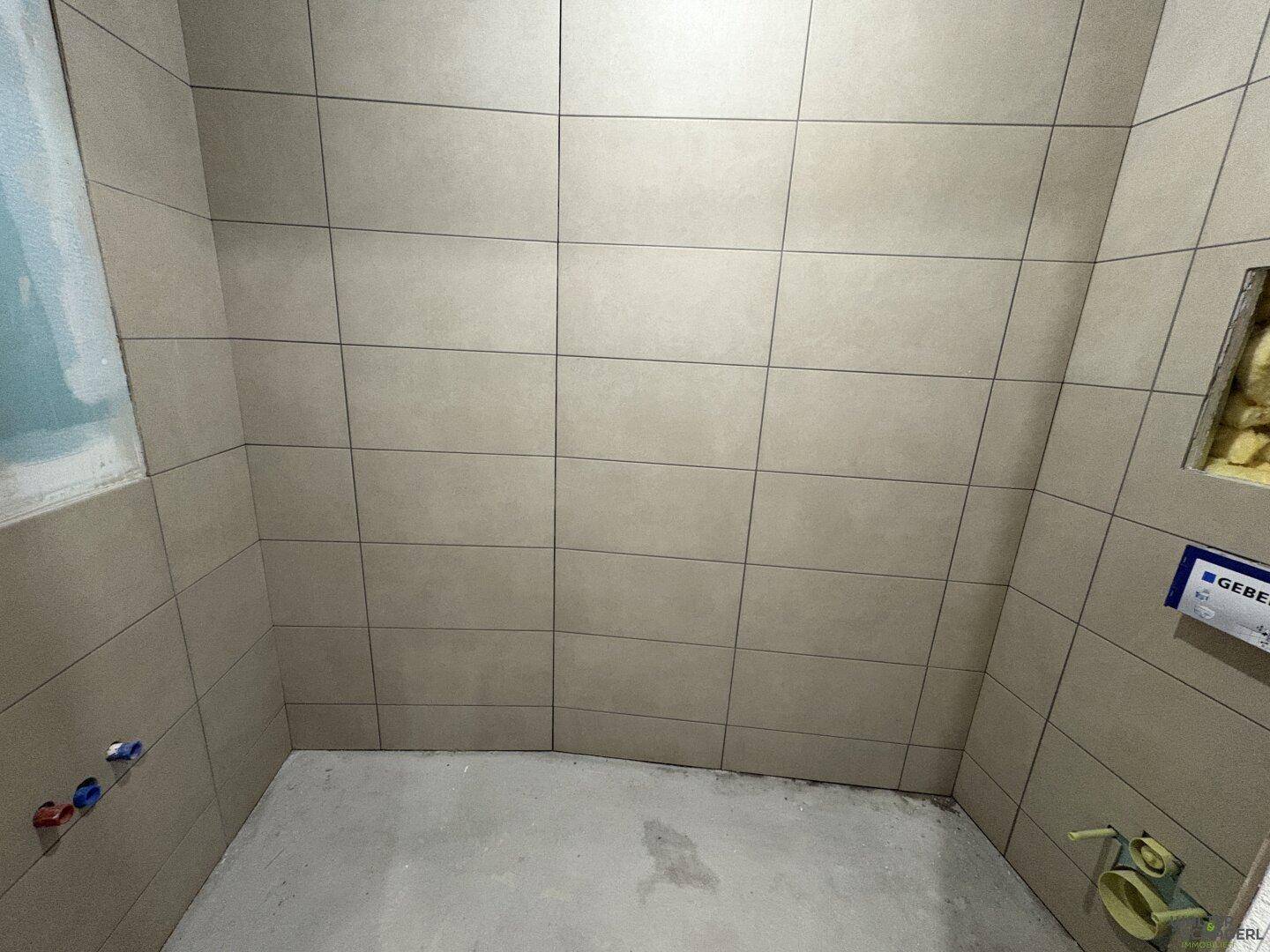 Badezimmer im Rohbau mit beigefarbenen Fliesen an den Wänden und vorbereiteten Anschlüssen.