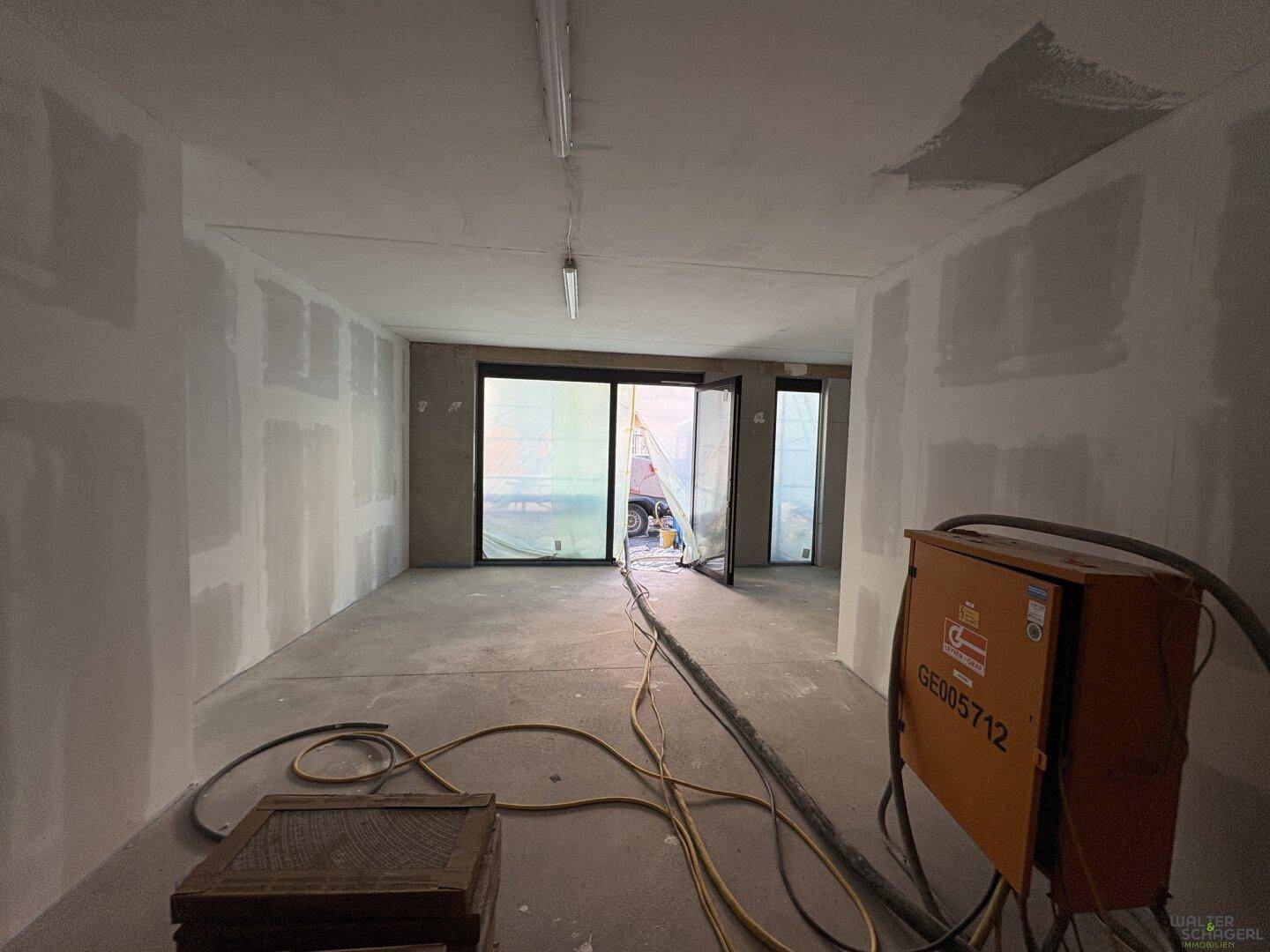 Rohbau-Innenraum mit großen Glasfronten, die Tageslicht hereinlassen, und Baugeräten auf dem Betonboden.