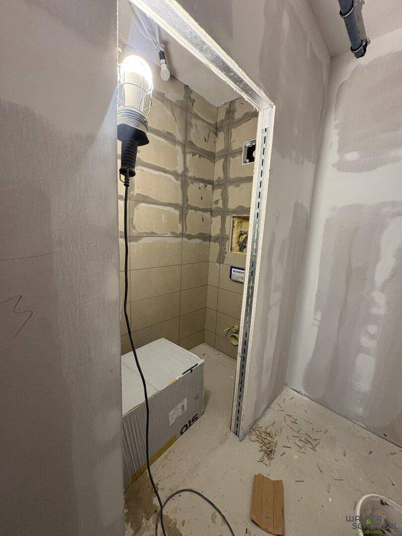 Kleiner Raum im Rohbau mit teilweise gefliesten Wänden und Baustellenbeleuchtung, vermutlich ein Badezimmer.