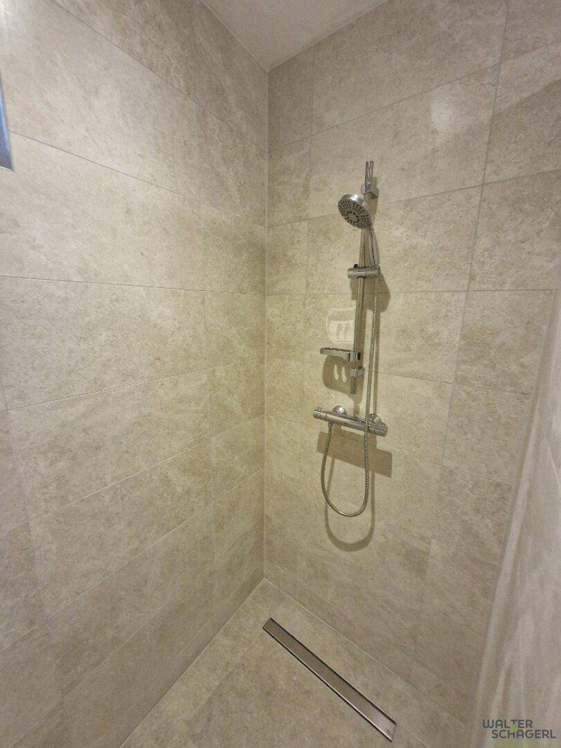 Detaillierte Ansicht einer modernen, gefliesten Dusche mit Duschkopf und Ablage.
