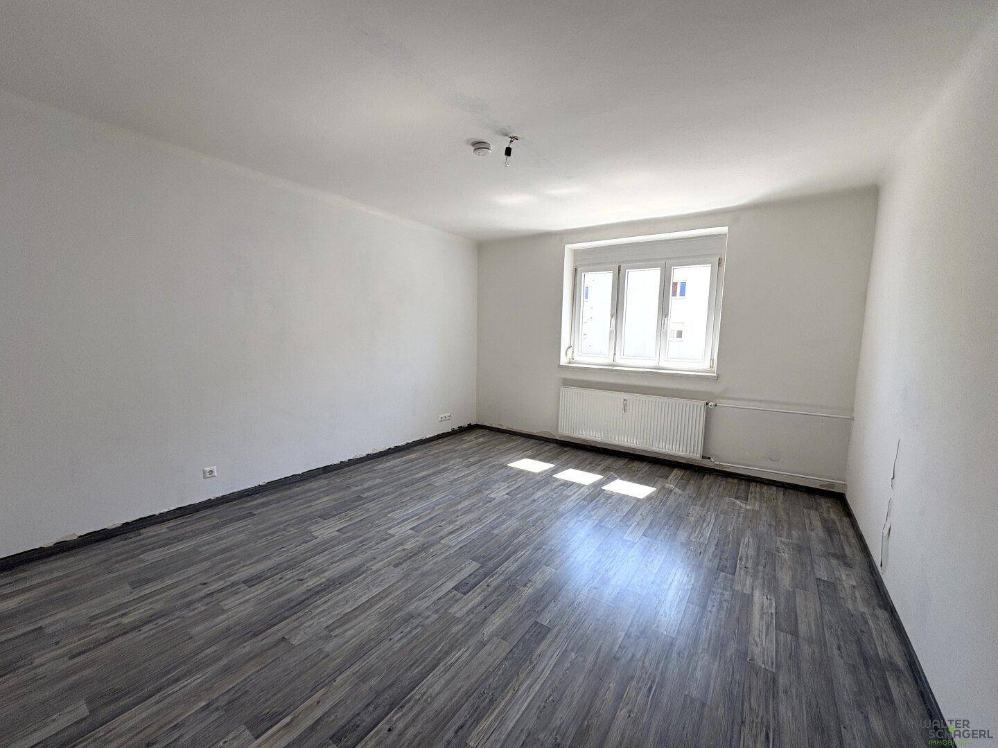 Geräumiger, heller Raum mit großem Fenster, Heizkörper und modernem Laminatboden.