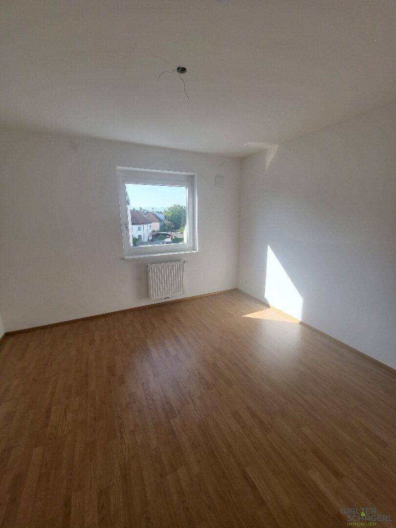 Helles, leeres Zimmer mit Holzfußboden, weiß gestrichenen Wänden und viel Tageslicht.