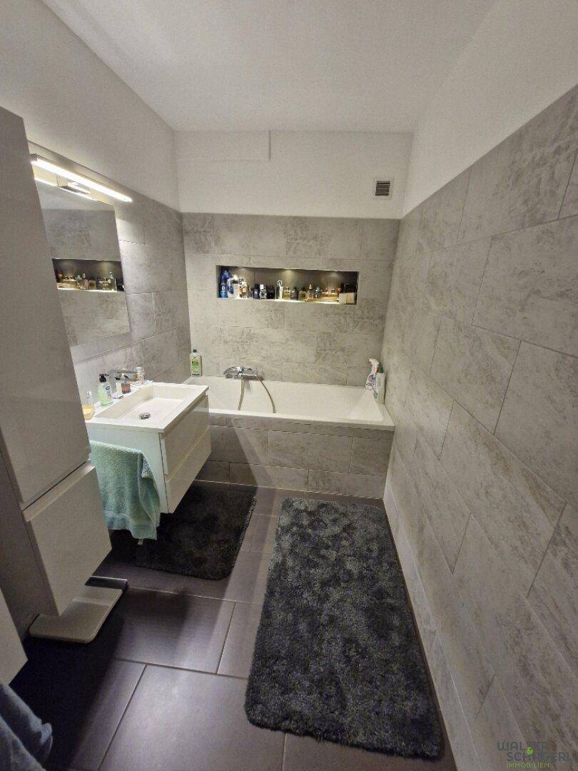 Modernes Badezimmer mit grauen Fliesen, Badewanne und integrierter Nische für Ablagen.
