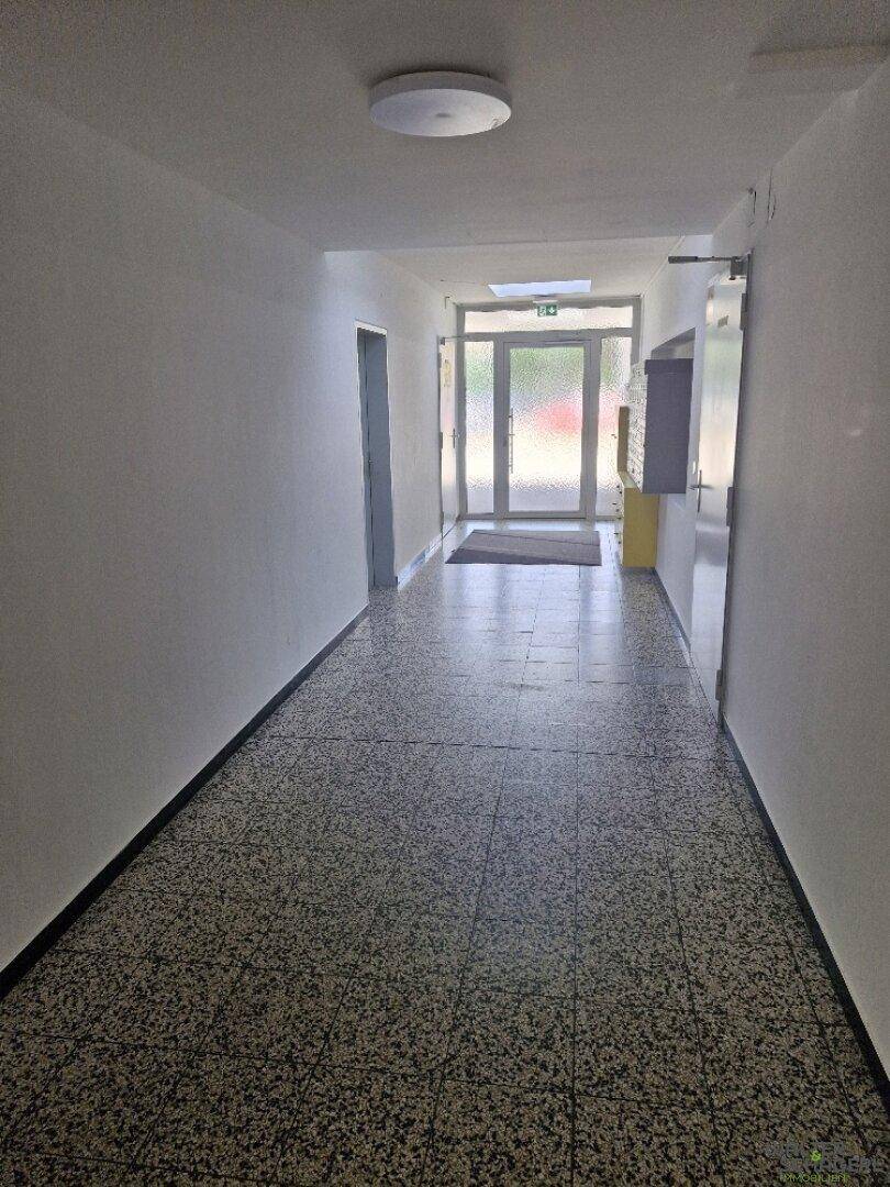 Langer Flur im Gebäude mit Terrazzoboden und hellem Eingangsbereich am Ende.