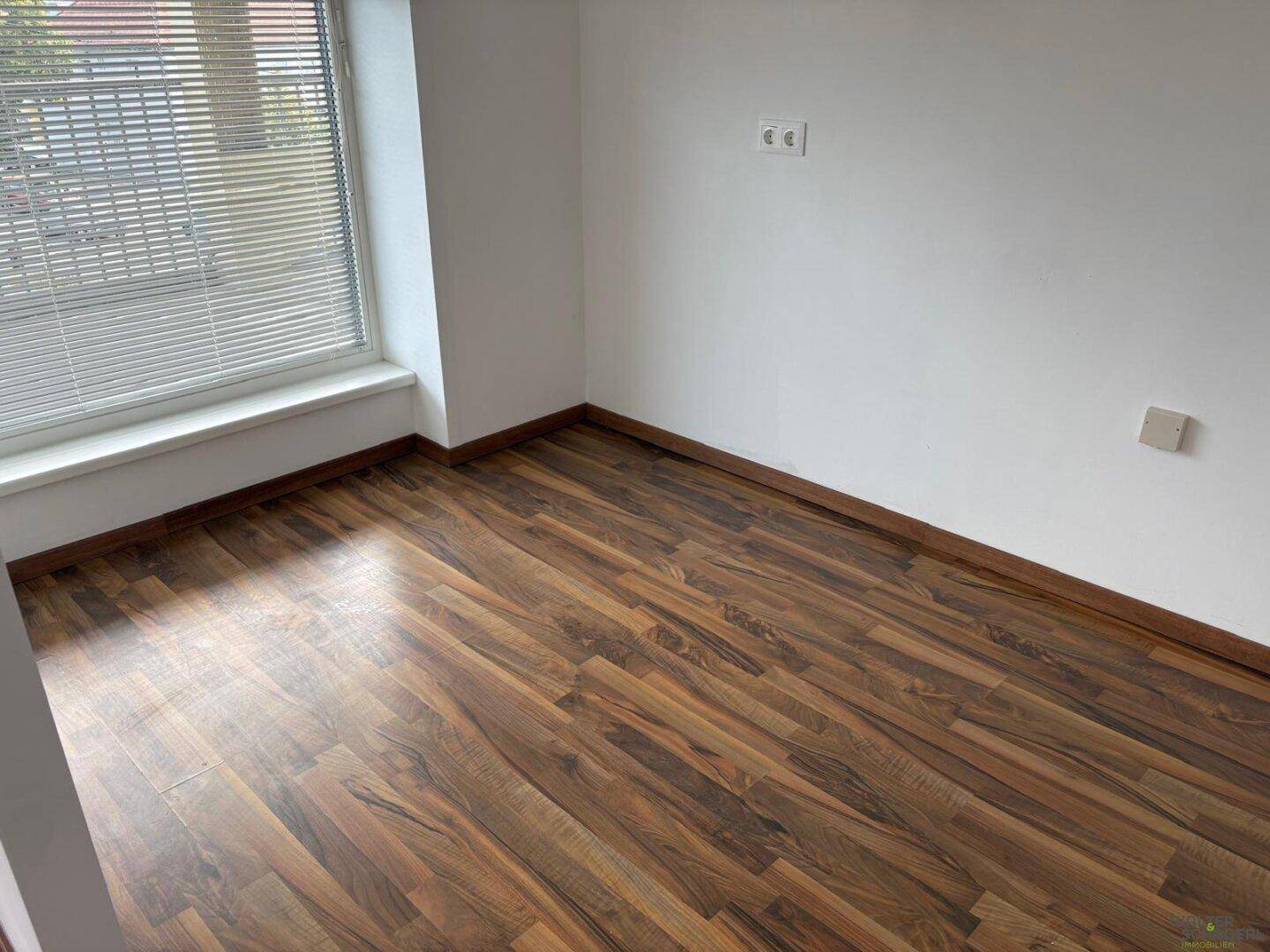 Leerer Raum mit Laminatboden und Fenster mit Jalousien, bereit zur individuellen Gestaltung.