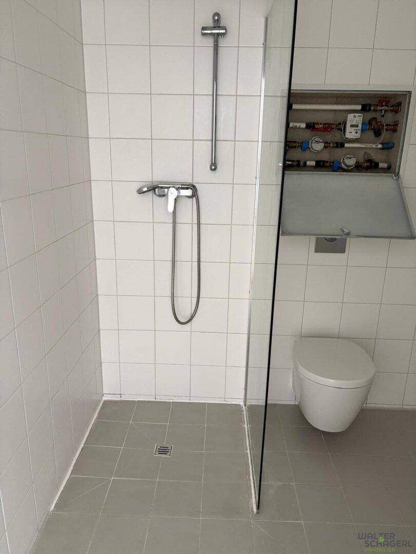 Duschbereich mit Glasabtrennung und wandhängendem WC in einem hell gefliesten Badezimmer.