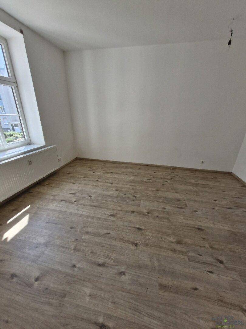 Leerer Raum mit Laminatboden, weißen Wänden und einem Fenster für natürliches Licht.