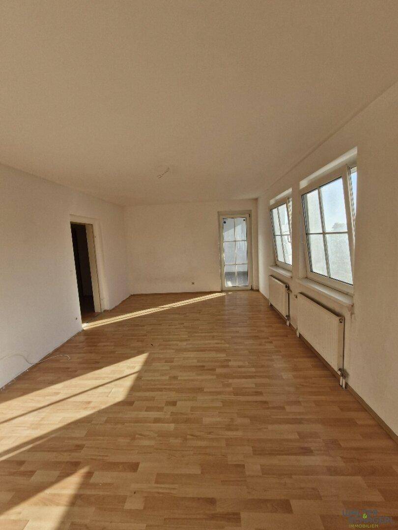 Geräumiges Zimmer mit Holzboden, weißen Wänden und mehreren Fenstern, die viel Licht hereinlassen.