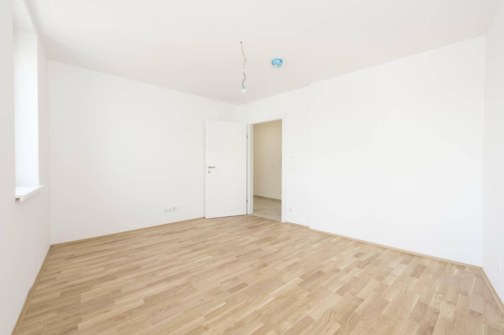 Helles Zimmer mit Parkettboden, weißen Wänden und einem Fenster für Tageslicht.