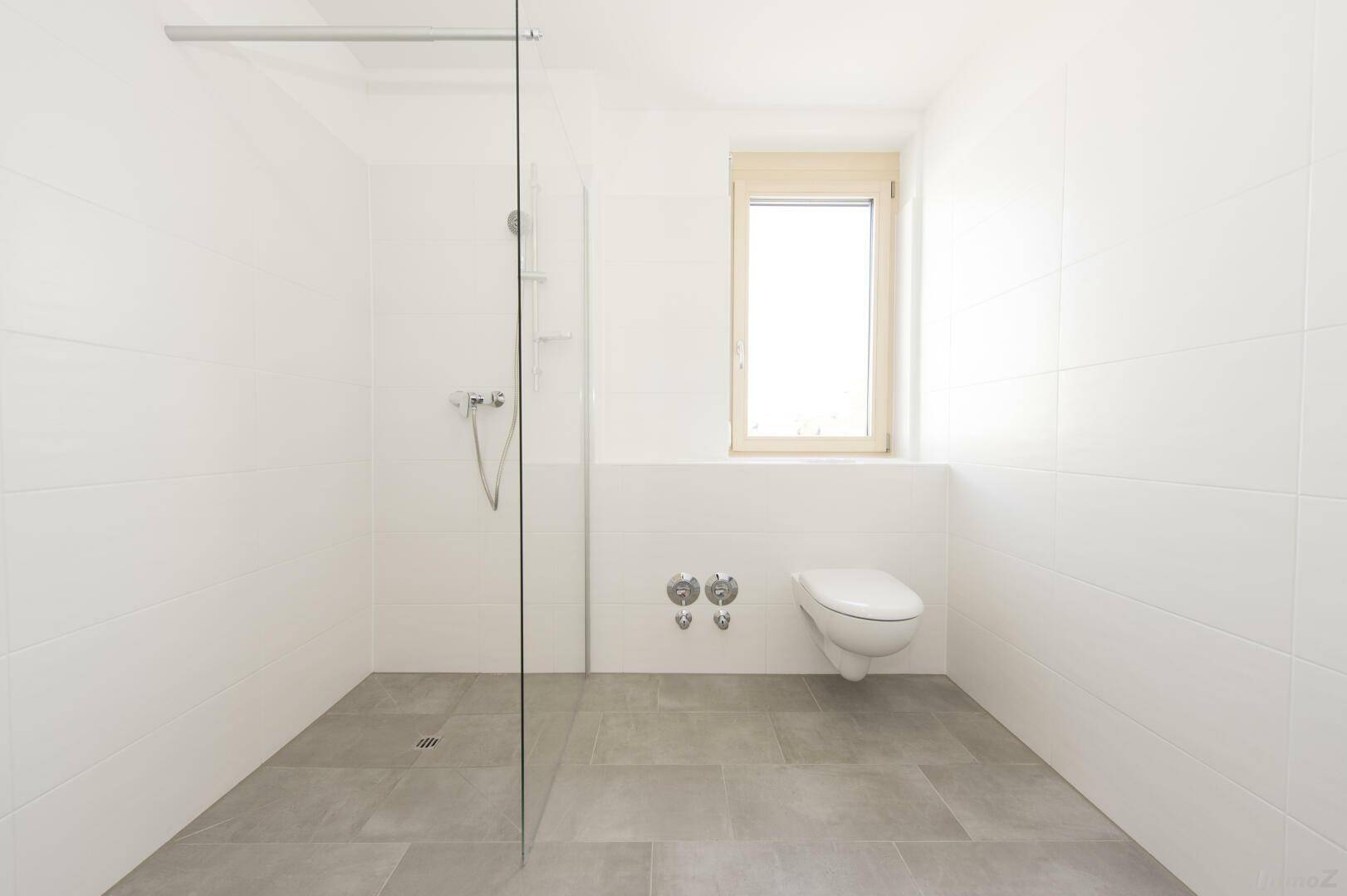 Helles Badezimmer mit großer ebenerdiger Dusche, WC und Fenster für natürliche Belüftung.