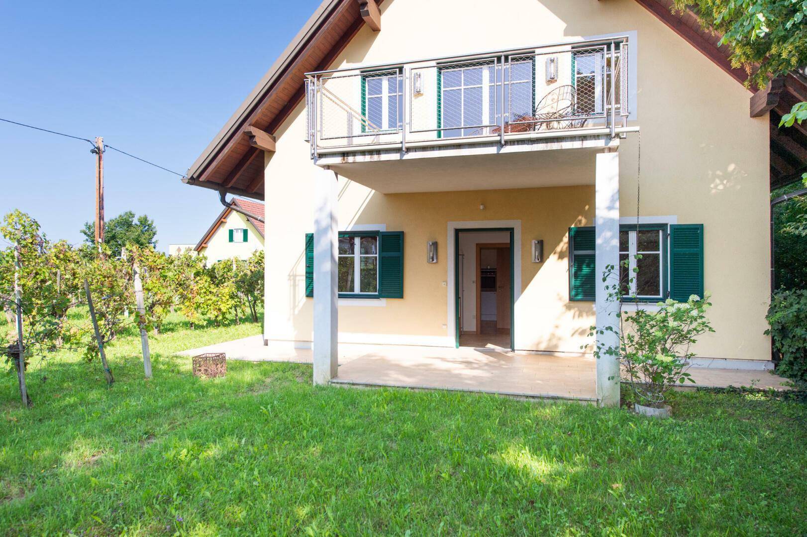 Hausfassade mit gepflasterter Terrasse und Rasenfläche, umgeben von Weinreben.