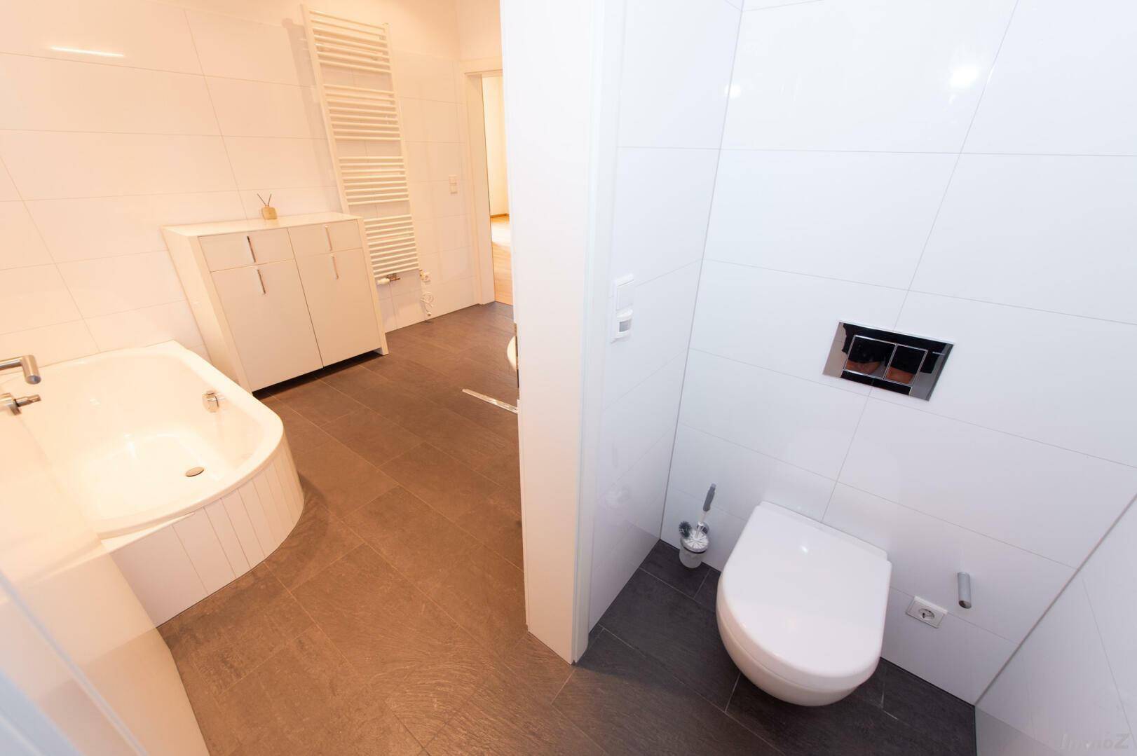 Helles Badezimmer mit Badewanne, wandhängendem WC und dunklem Fliesenboden.