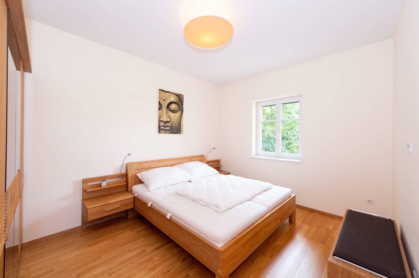 Schlafzimmer mit Holzbett, Nachttischen und Parkettboden, hell durch großes Fenster.