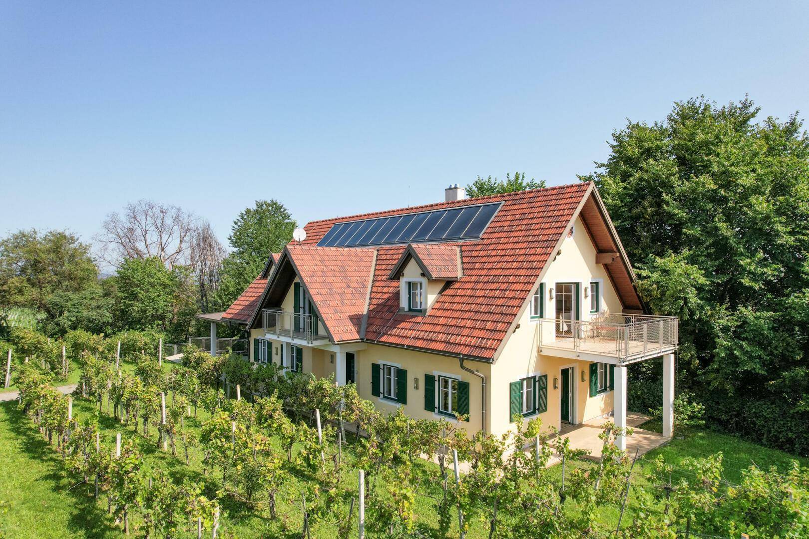 Ansicht des Hauses mit rotem Ziegeldach, grünen Fensterläden und Weinbergen im Vordergrund.