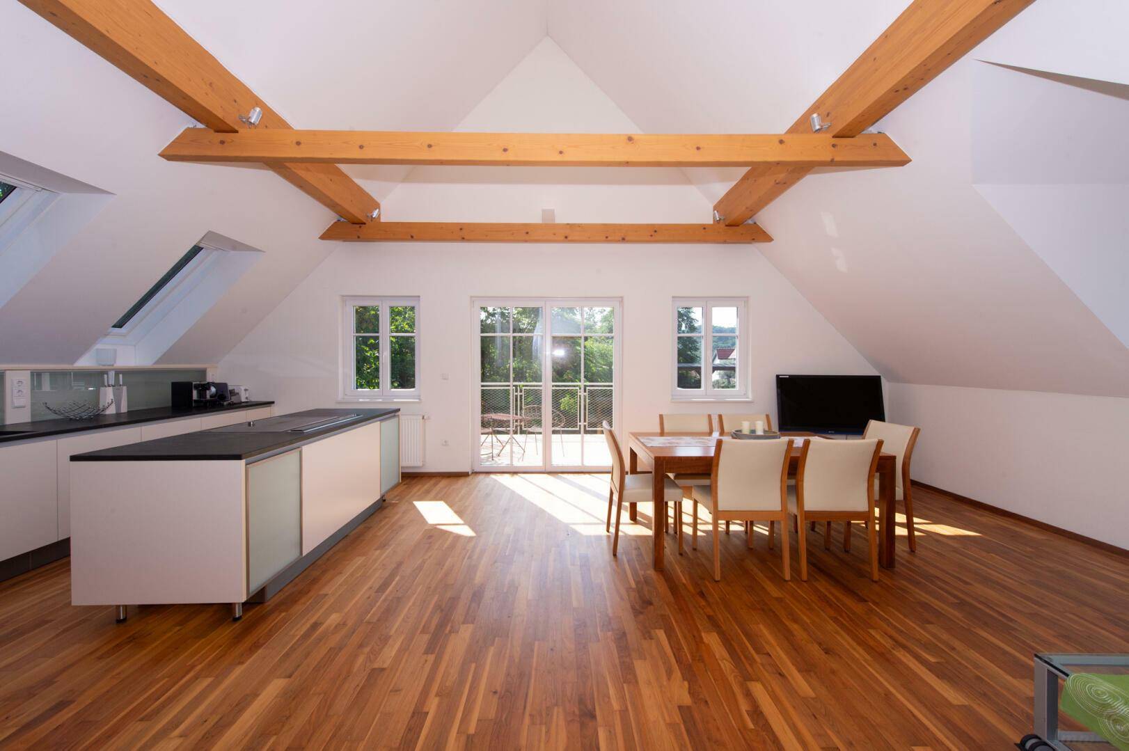 Offener Wohn-Essbereich im Dachgeschoss mit Holzbalken, Küche und Zugang zum Balkon.