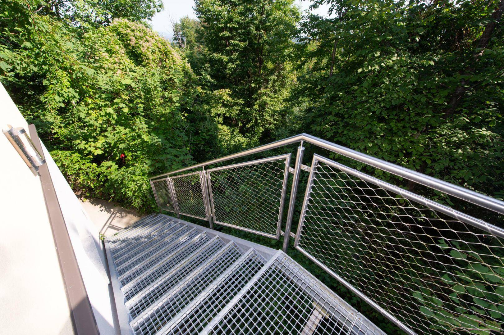 Außentreppe aus Metall mit Geländer, führt durch grüne Vegetation nach oben.
