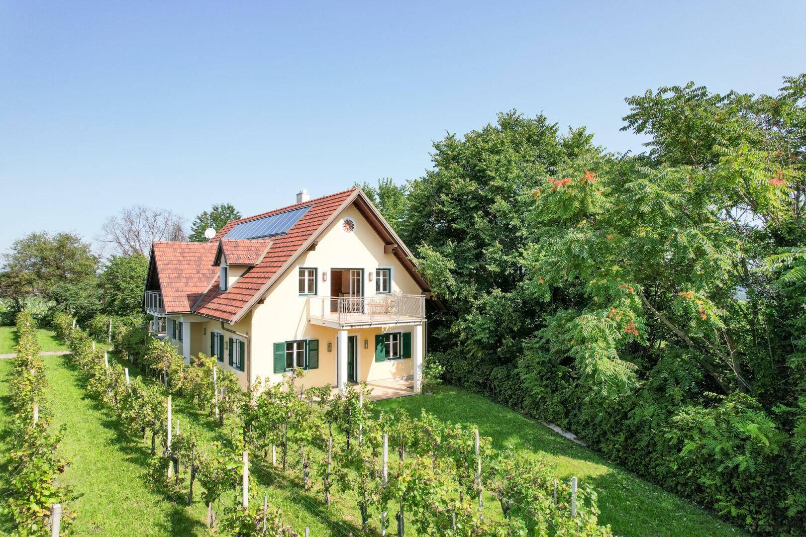 Seitenansicht des Hauses mit Balkon, umgeben von üppiger Vegetation und Weinreben.