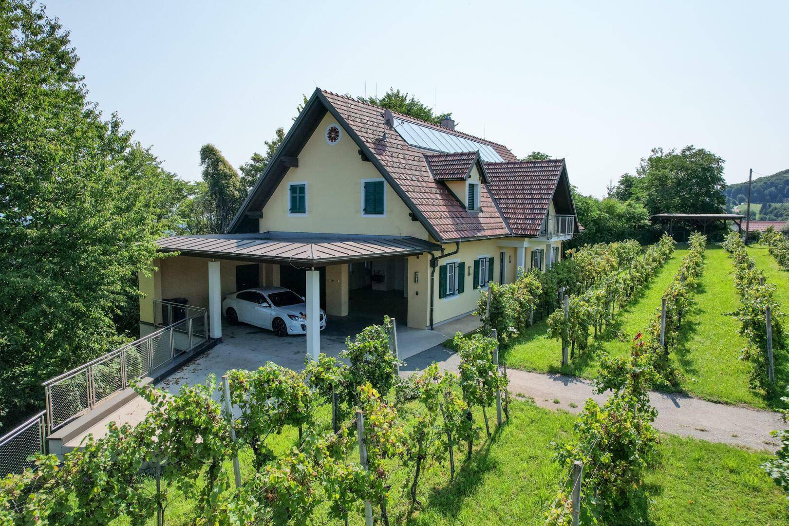Haus mit Carport und Solaranlage, umgeben von gepflegten Weinbergen unter blauem Himmel.