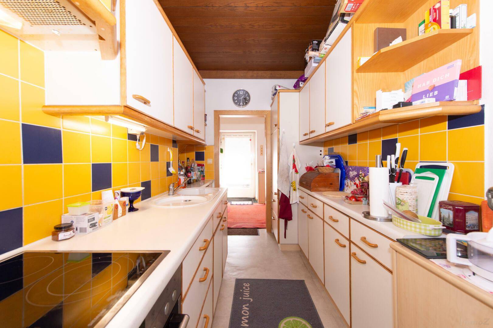Schmale Küche mit gelb-blauen Fliesen, weißen Schränken und Holzdecke.