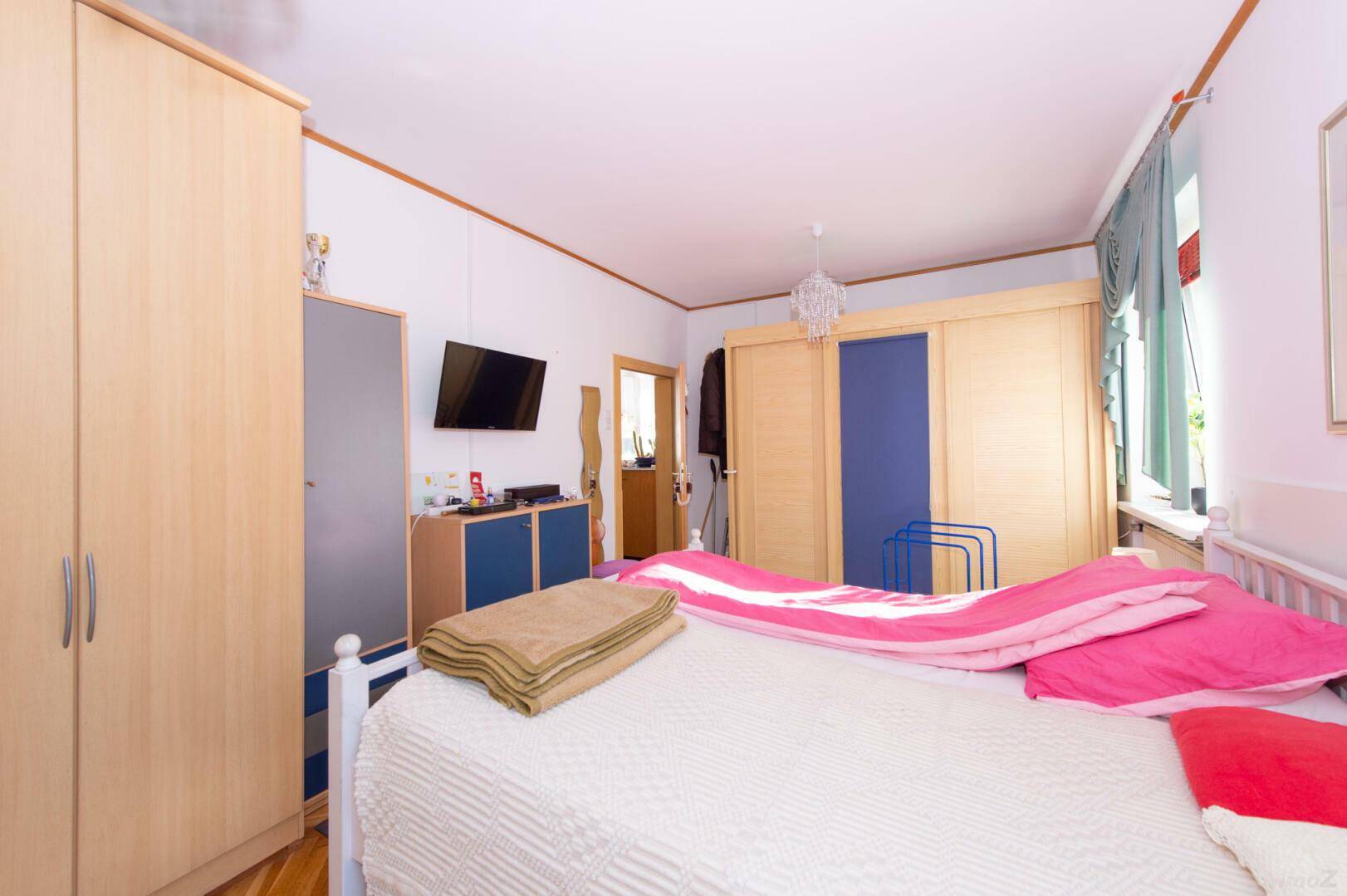 Schlafzimmer mit Holzboden, hellem Bett und Fernseher an der Wand.