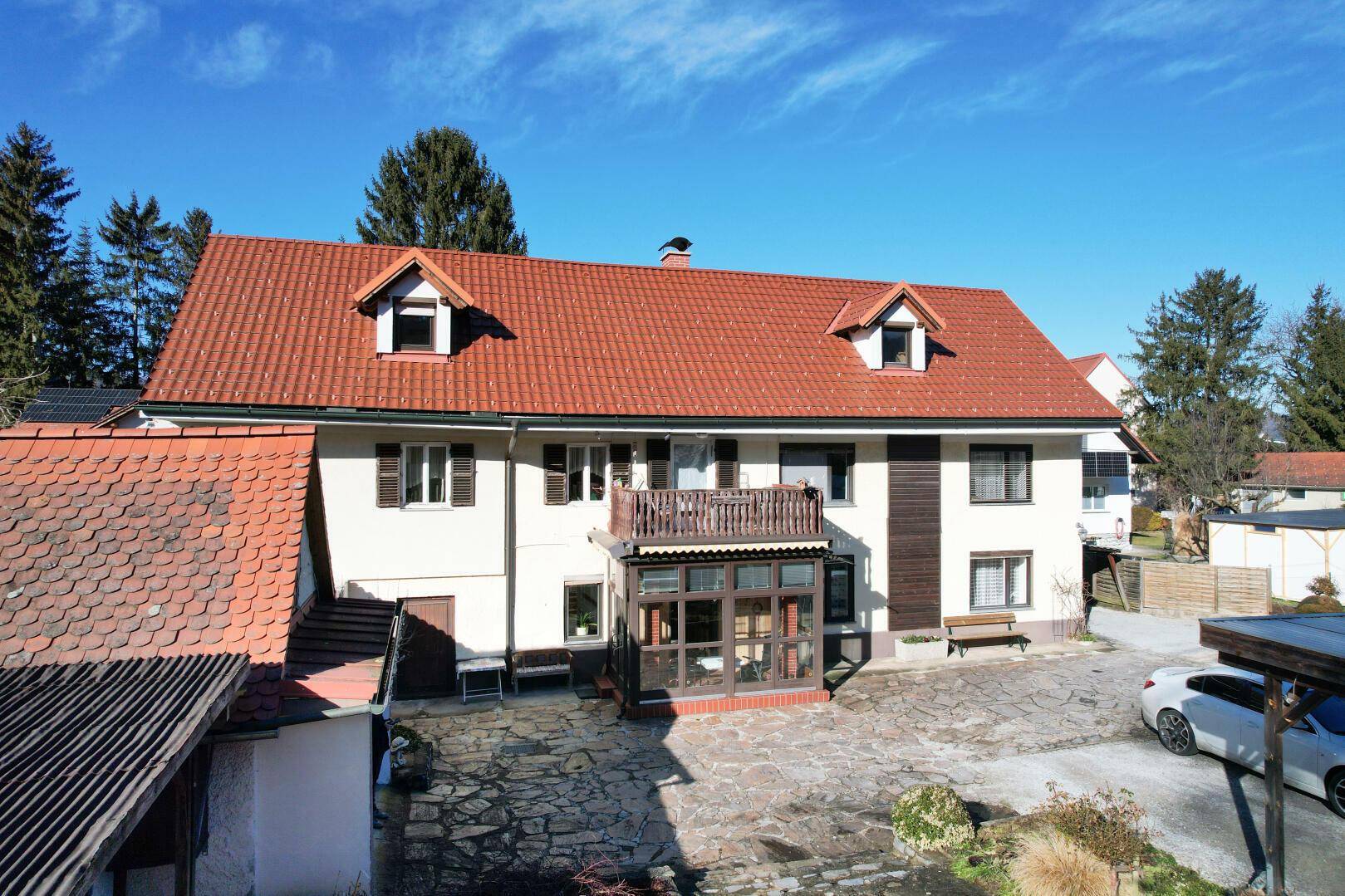 Die Vorderansicht des Hauses mit rotem Ziegeldach, Wintergarten und gepflasterter Fläche.