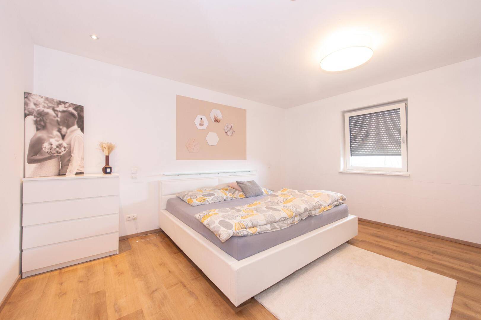 Helles Schlafzimmer mit modernem Bett, Holzboden und Fenster mit Rollladen.