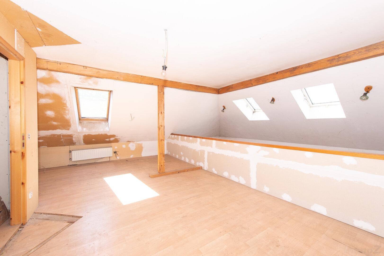 Teilweise renovierter Dachbodenraum mit Holzboden, sichtbaren Balken und Dachfenstern.
