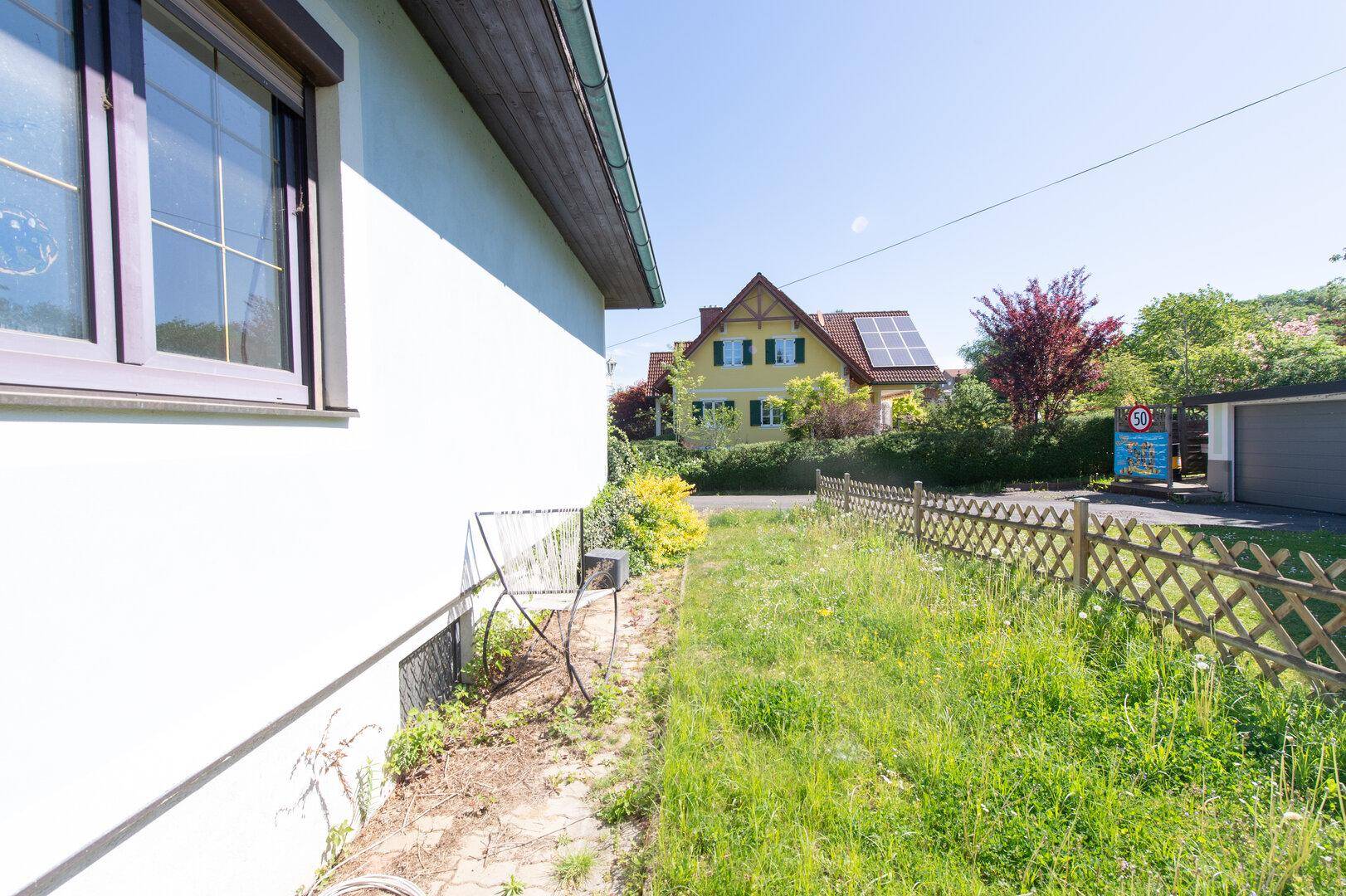 Seitenansicht eines Hauses mit grünem Rasen, Zaun und Nachbargebäuden im Hintergrund.
