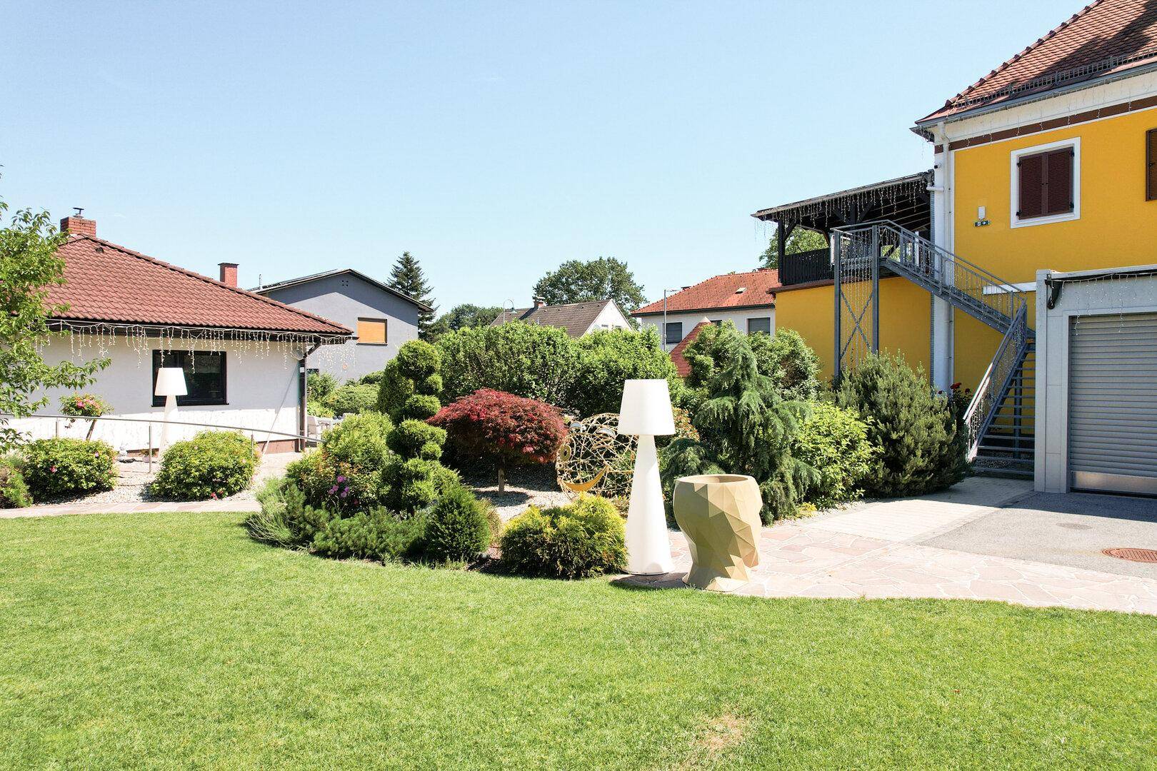 Gelbes Gebäude mit Außentreppe und Garage, umgeben von einem grünen Garten mit dekorativen Elementen.