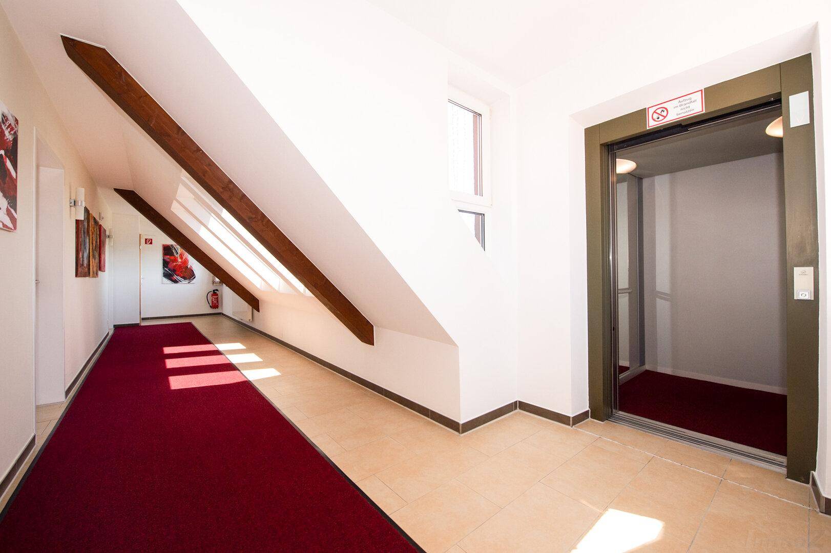 Heller Flur im Dachgeschoss mit Teppichboden, Dachschrägen und einem Aufzug.