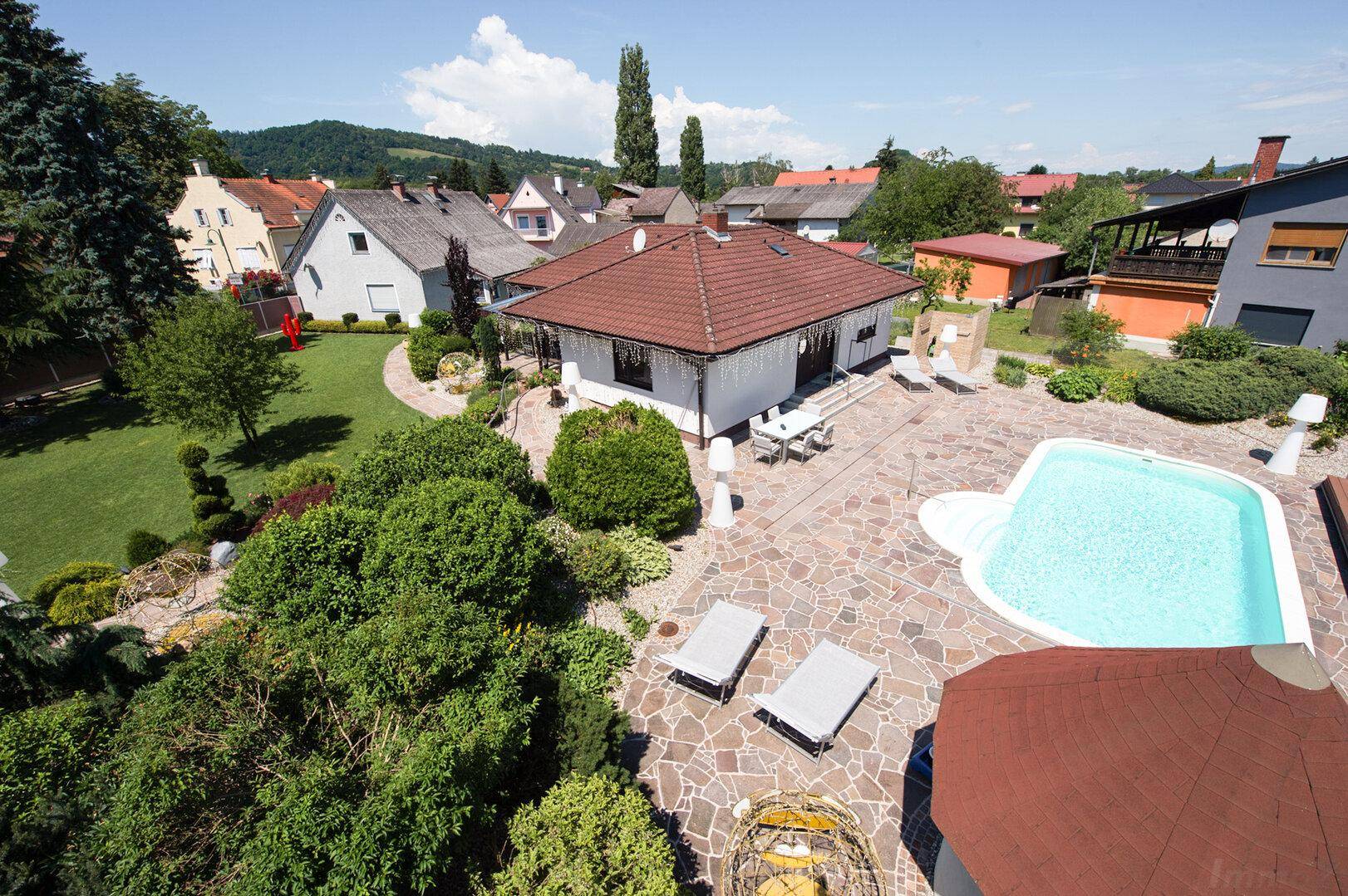 Luftaufnahme eines Anwesens mit großem Garten, Swimmingpool und gepflasterter Terrasse unter blauem Himmel.