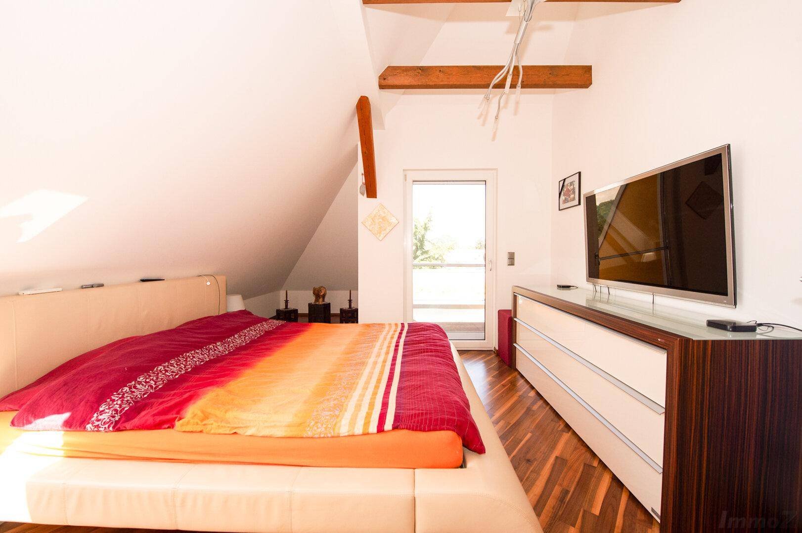 Schlafzimmer im Dachgeschoss mit Holzboden, TV und Zugang zum Balkon.