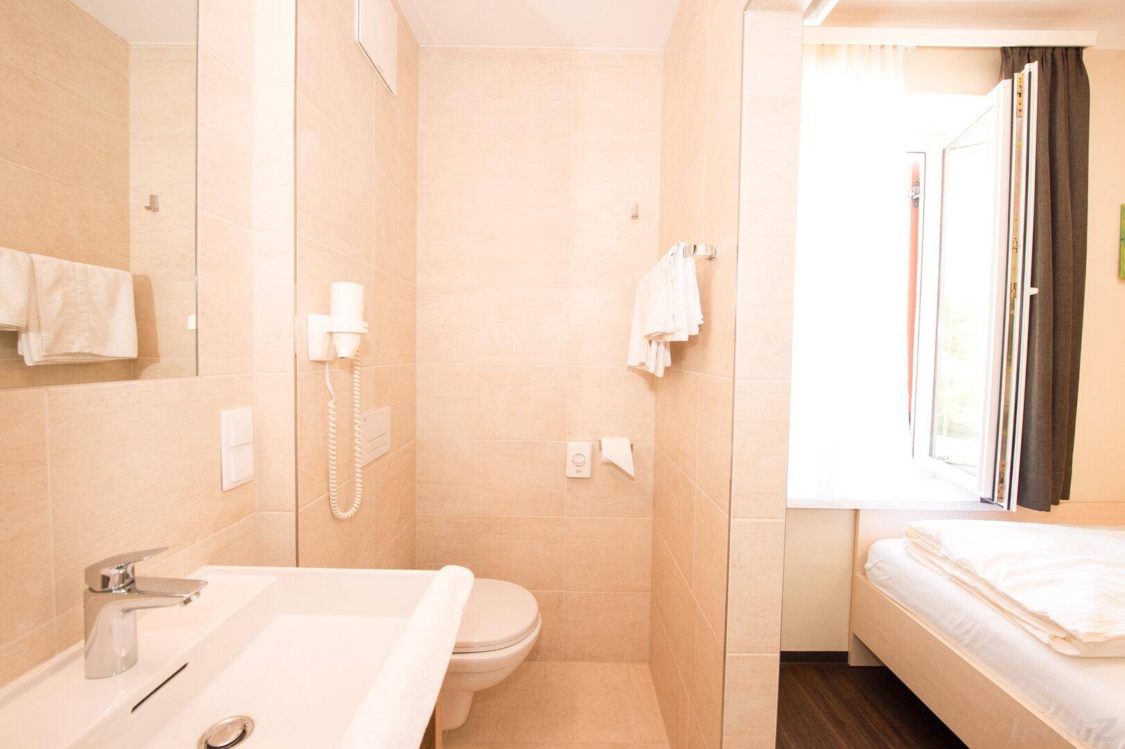 Helles Badezimmer mit Fenster, Waschbecken und Toilette, angrenzend an ein Schlafzimmer.