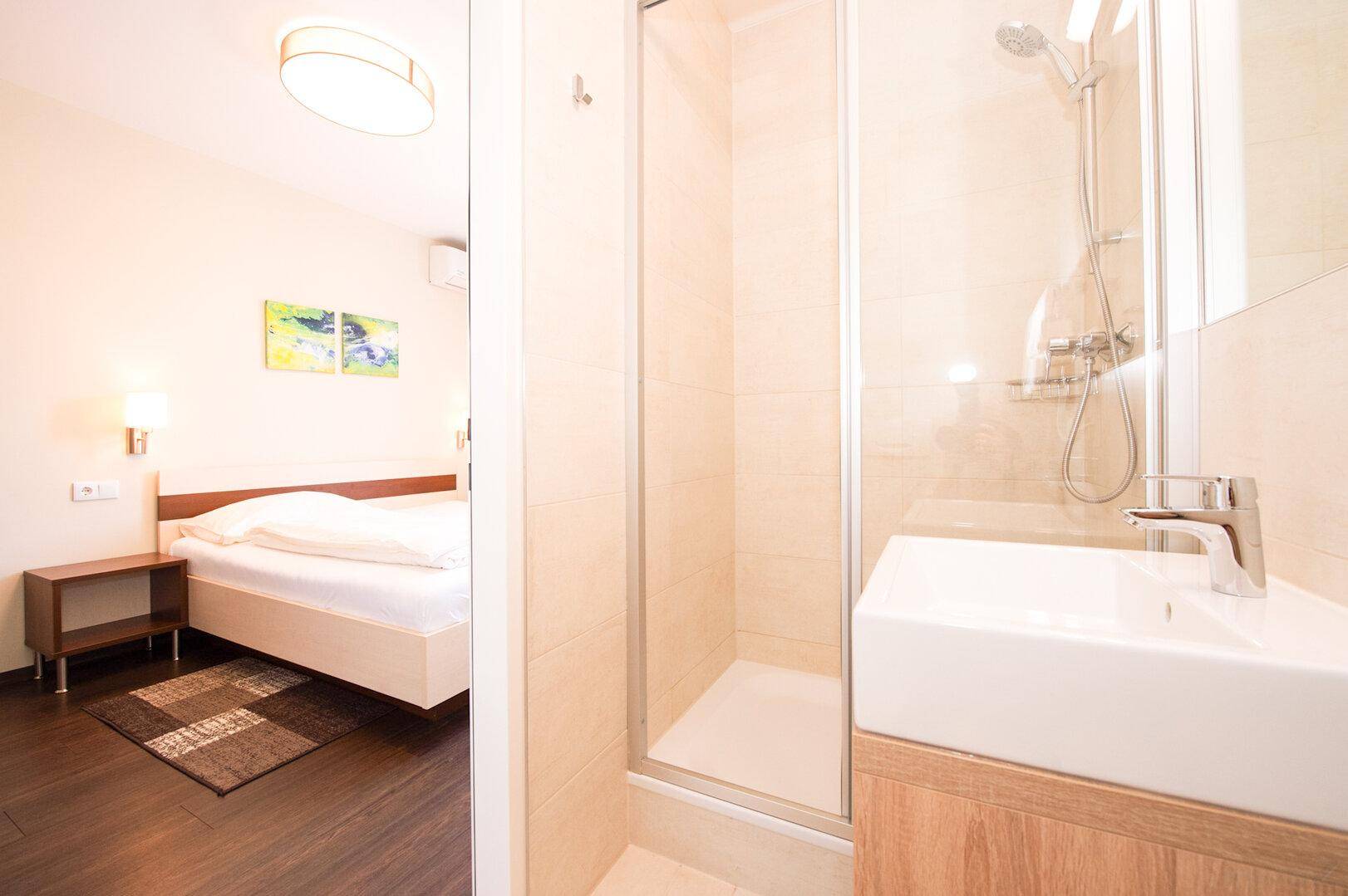 Kompaktes Badezimmer mit Duschkabine, Waschbecken und Blick in ein angrenzendes Schlafzimmer.