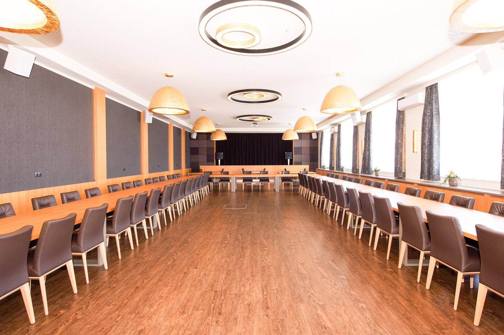 Großer Konferenzraum mit langen Tischen, vielen Stühlen und modernen Deckenleuchten.