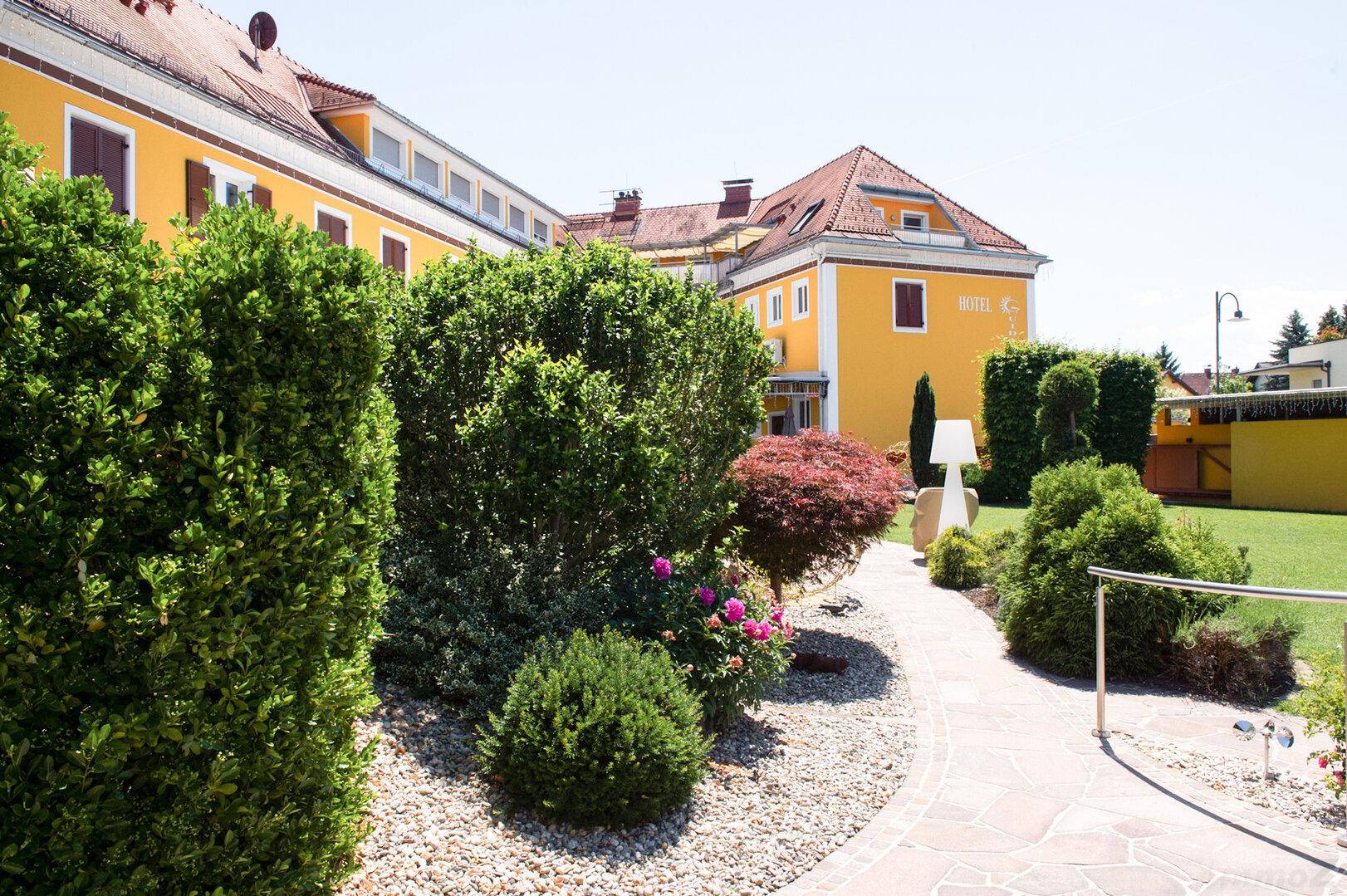 Geschwungener Gartenweg mit Kies und gepflegten Hecken führt zum gelben Hotelgebäude unter blauem Himmel.