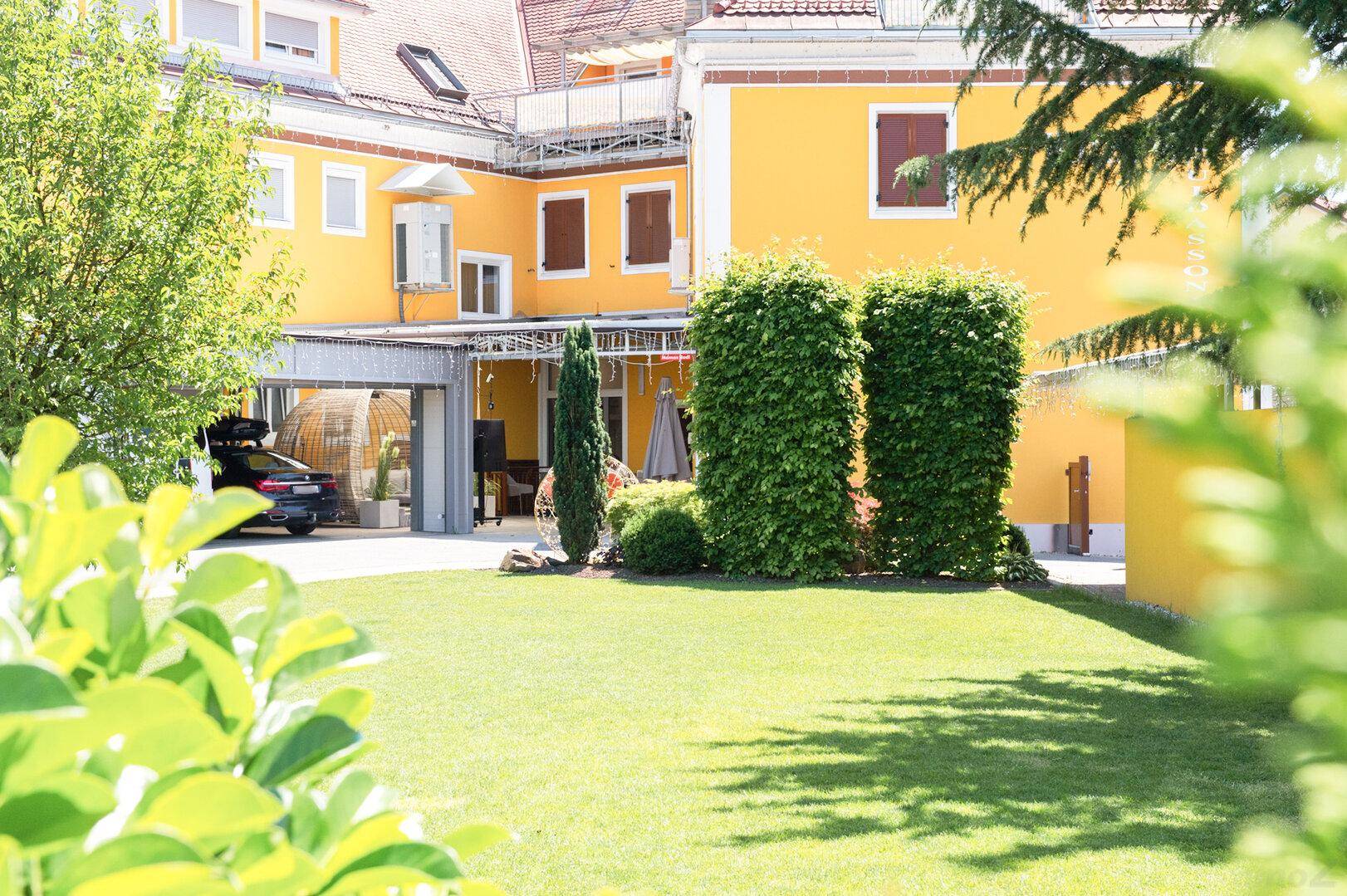 Grüner Rasenbereich mit Bäumen und Hecken, im Hintergrund das gelbe Hotelgebäude mit Carport.