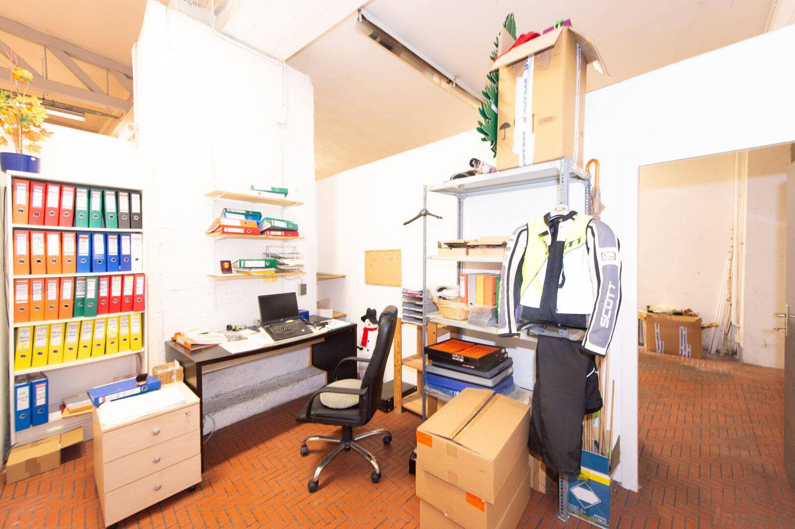 Kleines Büro mit Schreibtisch, Regal und Aktenordnern, funktional eingerichtet.