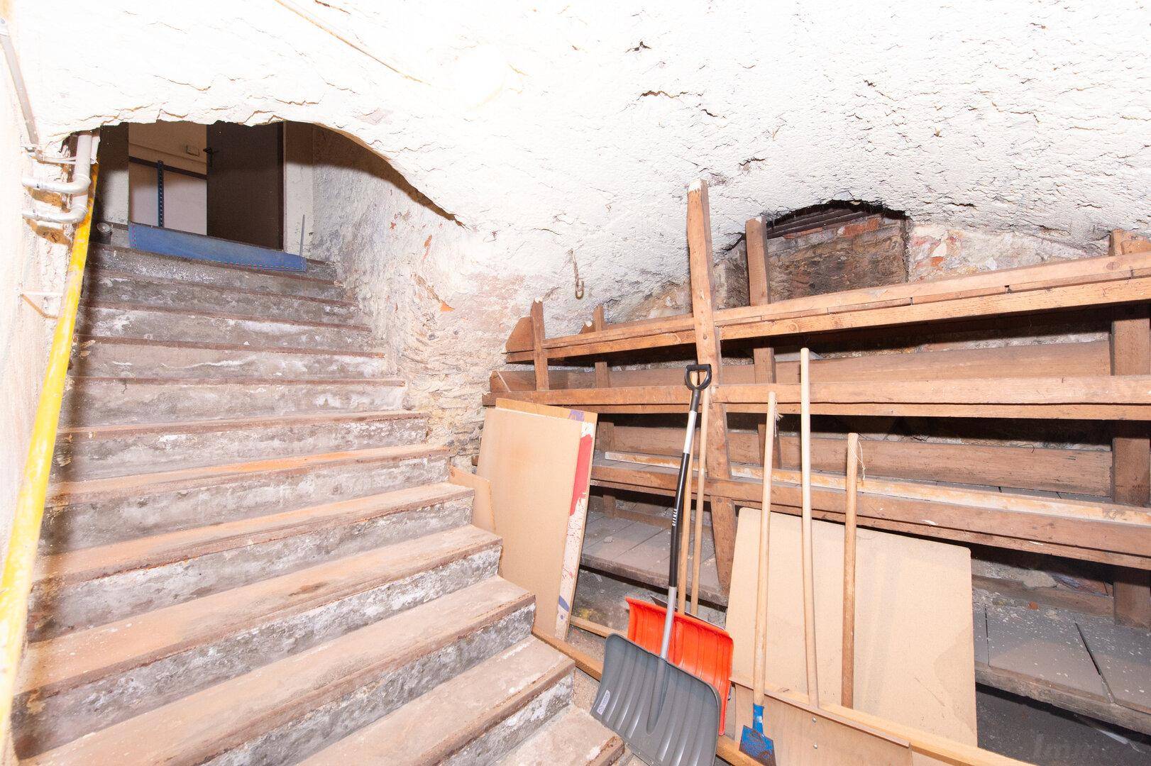 Kellertreppe führt hinunter zu einem Lagerbereich mit einfachen Holzregalen.