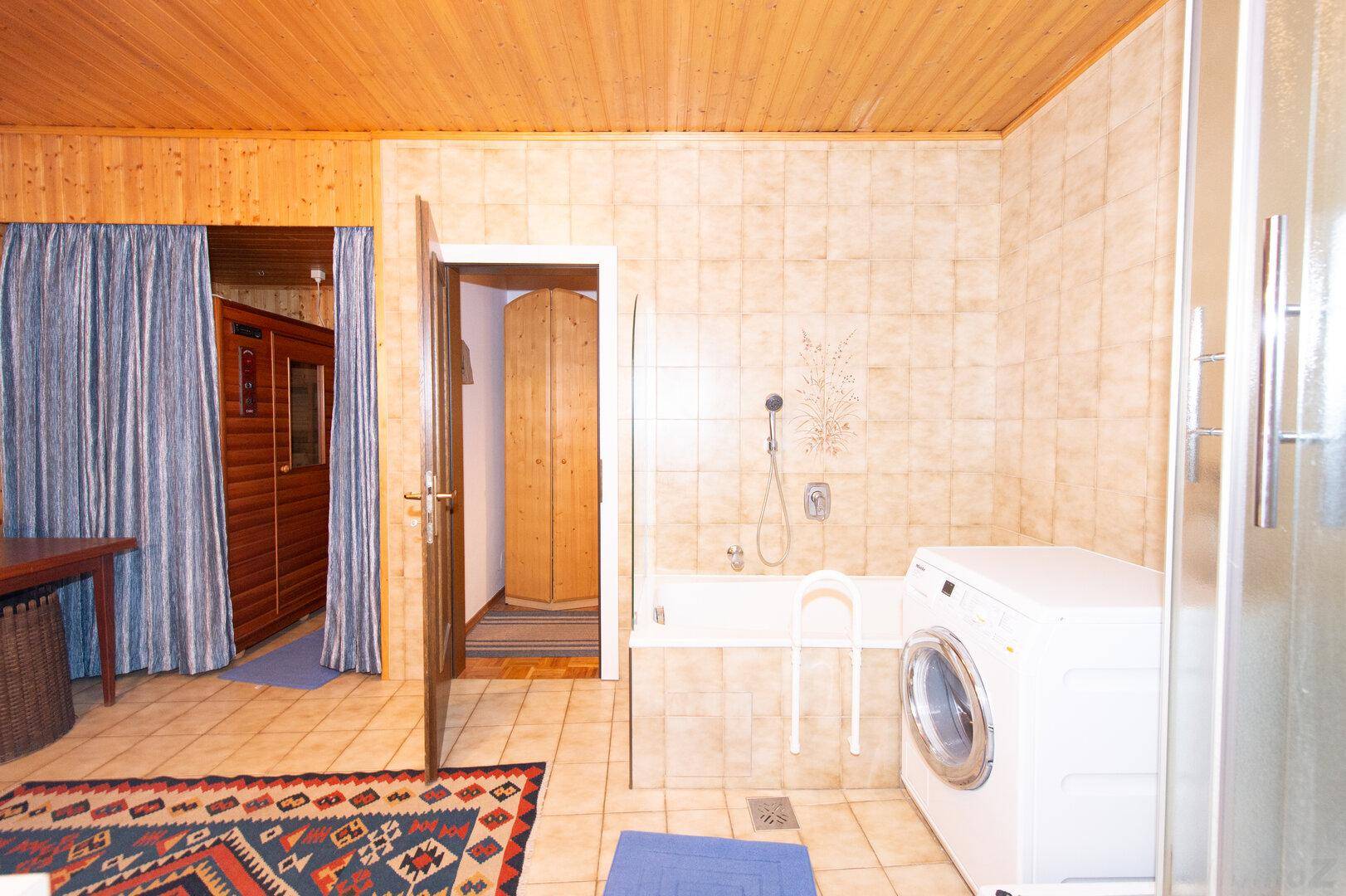 Badezimmer mit Waschmaschine, Duschkabine und Badewanne, umgeben von beigefarbenen Wandfliesen.