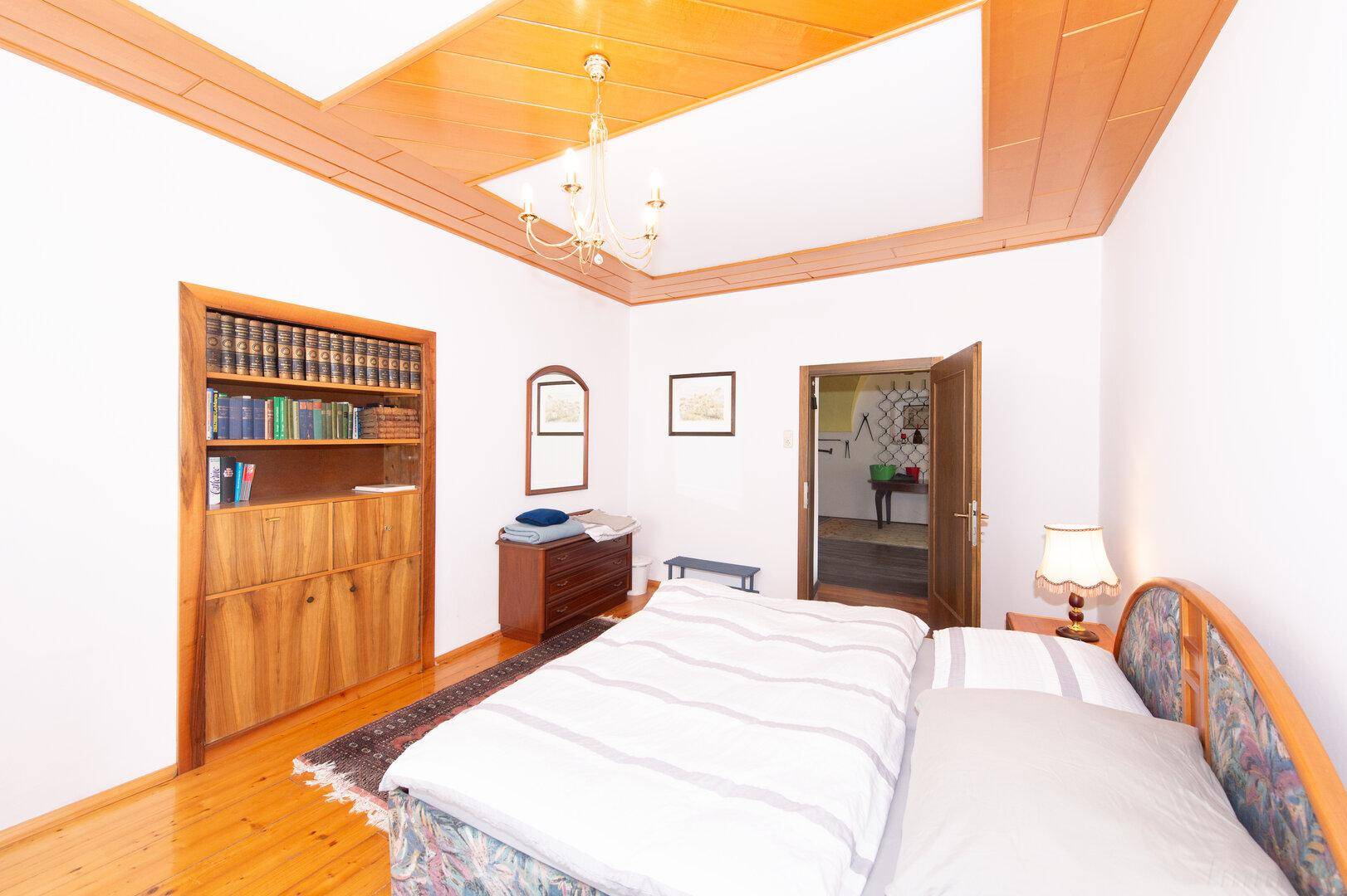 Schlafzimmer mit Bett, Bücherregal und einem Spiegel, ergänzt durch eine Holzdecke.