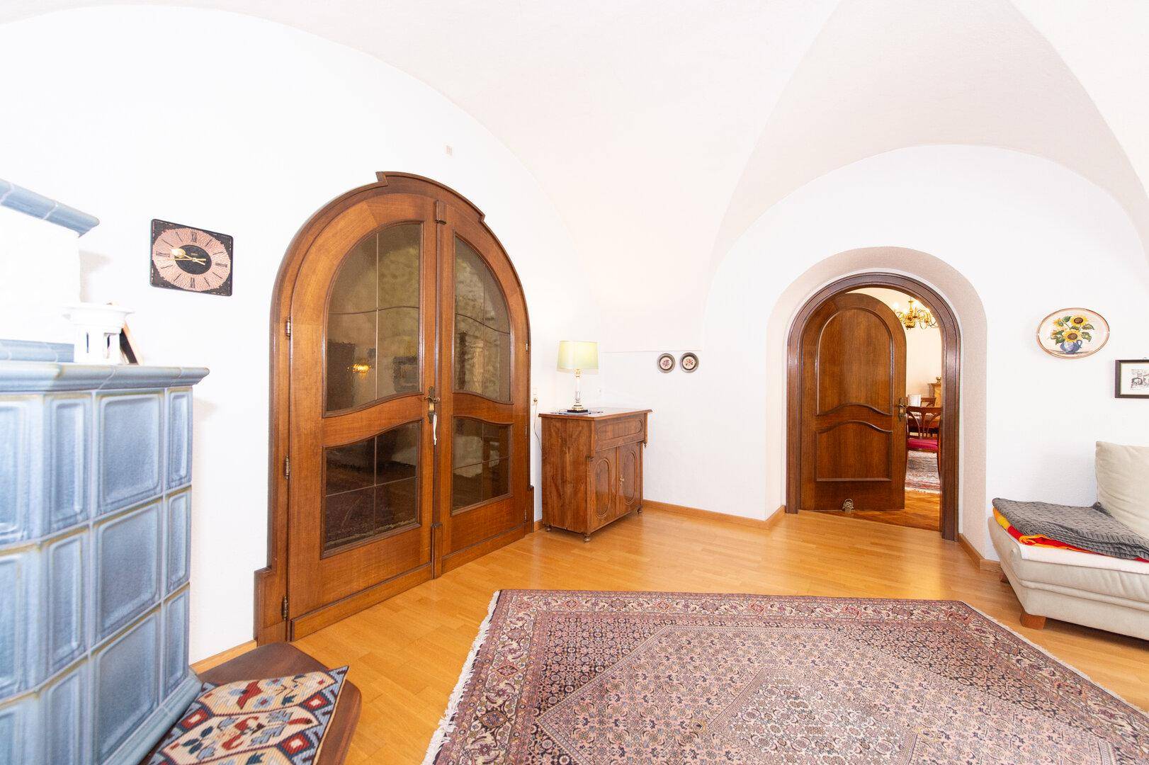Wohnbereich mit Holzboden, gewölbter Decke, Kachelofen und großen Holztüren, sehr stilvoll.